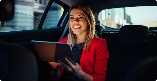 Woman in a red jacket using a tablet and smiling while seated in the back of a car at night.