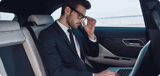 Man in business attire working on his laptop in the rear seat of a black luxury vehicle.