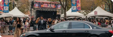 Black car parked outside a busy event at Made in America Fest, with crowds and white tents in view.
